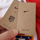 Camisola principal do Atlético de Madrid 2025/26 da Nike, com riscas vermelhas e brancas, gola branca, escudo com contorno amarelo e logótipo azul royal.