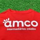Camisola vermelha com estampado branco da marca amco, intermediaríos crédito, em fundo de grama sintética.