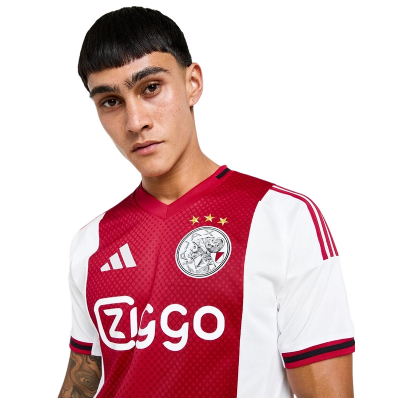 Camisola principal do Ajax 2025/26 Adidas em branco e vermelho com faixa central em padrão gradiente, gola em V e detalhes icónicos da cidade de Amesterdão.