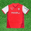 Camisola SC Braga home 24/25 Vermelho na grama, com detalhes em branco e logotipos da Puma e patrocinadores.