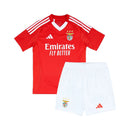 Conjunto infantil titular do Benfica 2024/25 com camisola vermelha, gola em V branca, chevrons em embossing e calção branco. Modelo clássico e moderno para jovens benfiquistas.