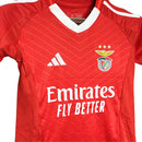 Conjunto infantil titular do Benfica 2024/25 com camisola vermelha, gola em V branca, chevrons em embossing e calção branco. Modelo clássico e moderno para jovens benfiquistas.