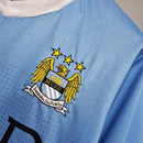 Emblema do Manchester City em camiseta retro azul 11/12, destacando as três estrelas e detalhes em bordado.