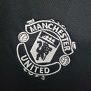 Emblema bordado do uniforme Manchester United, com detalhes em branco sobre fundo preto.